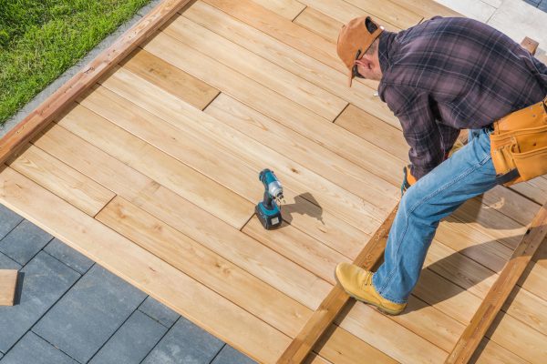Caucasian Men in His 40s Building Wooden Deck on His Backyard Attaching Wood Board To Each Other Using Cordless Drivers and Screws. Construction Industry Theme.